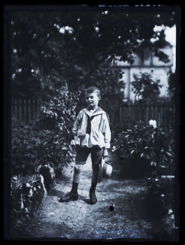 James Hertig enfant, en costume marin, tenant son chapeau à la main, posant dans un jardin