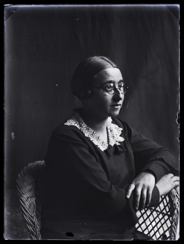 Portrait de femme portant des lunettes, vue de 3/4 droit, assise dans un fauteuil en osier, les mains croisées