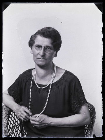 Portrait d'une femme portant des lunettes et un sautoir en perles, assise dans un fauteuil en osier