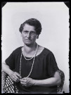 Portrait d'une femme portant des lunettes et un sautoir en perles, assise dans un fauteuil en osier